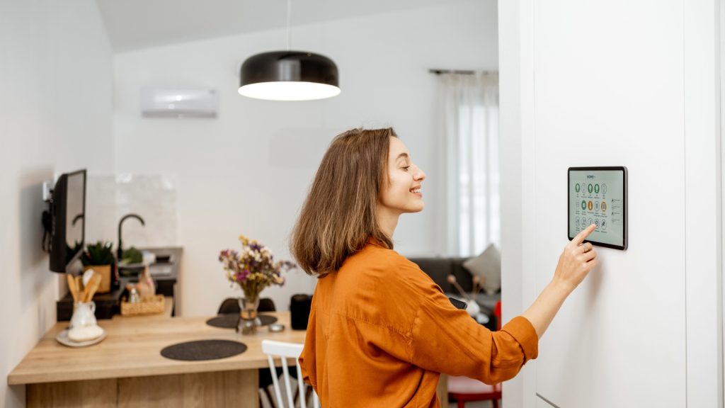 Frau mit schulterlangem Haar und orangefarbener Bluse bedient Wandtablet in modern eingerichteter Küche mit Holzarbeitsplatte und Blumenstrauß auf dem Tisch
